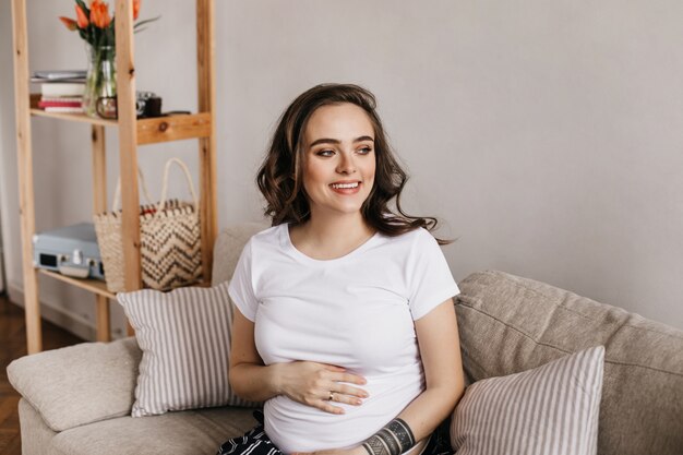 Cheerful young pregnant woman in white tee smiles and gently touches belly