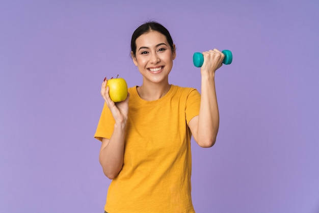 Cheerful young pretty sportswoman standing isolated over violet wall, showing yellow apple while lifting weights