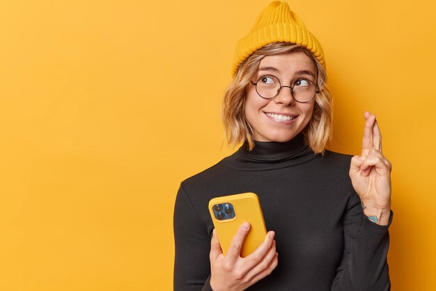 Cheerful young pretty woman keeps fingers crossed makes wish holds smartphone waits for dreams come true wears hat black turtleneck round spectacles isolated over yellow background copy space