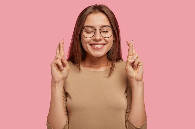 ポジティブな表情の陽気な若いかなり若い女性は、指を交差させ、夢の実現を望み、願い事をし、丸いメガネとカジュアルなセーターを着て、ピンクの壁に隔離されています。