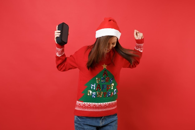 쾌활한 젊은 산타 소녀 춤과 점프, 빨간색 배경에 고립 된 휴대용 무선 블루투스 음악 스피커를 들고. 새해 복 많이 받으세요 2019 축 하 휴일 파티 개념입니다. 복사 공간을 비웃습니다.