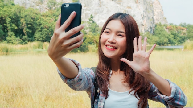 Cheerful young traveler Asian lady with backpack selfie at mountain lake. Korean girl happy using mobile phone taking selfie enjoy holidays on hiking adventure. Lifestyle travel and relax concept.