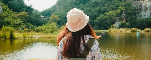 Cheerful young traveler Asian lady with backpack walking at the mountain lake. Korean teen girl enjoy her holidays adventure feeling happy freedom. Lifestyle travel and relax in free time concept.