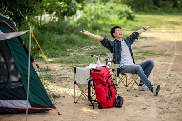 夏休みのキャンプ旅行中にコーヒーセットと淹れたてのコーヒーグラインダーを作る森のテントの前に座っている陽気な若い旅行者の男