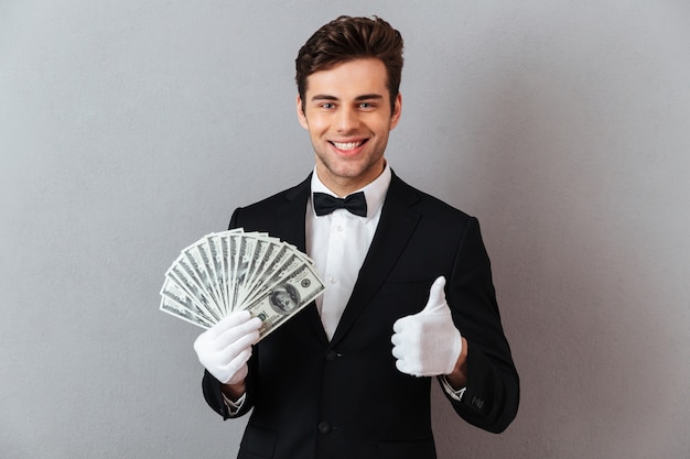 Веселый молодой официант показывает палец вверх держа деньги.