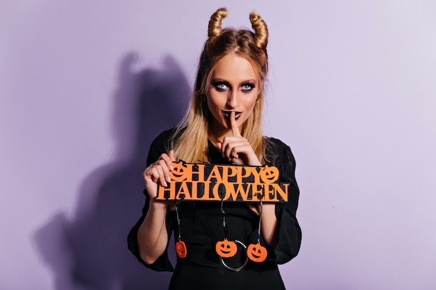 Cheerful young witch posing on purple wall with cute smile. wonderful blonde lady enjoying halloween party.