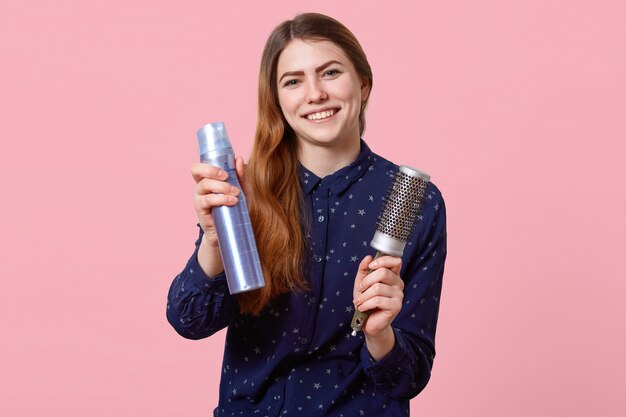 Cheerful young woman advertises new hairspray, holds comb, wants to change image, dressed in stylish shirt, poses over pink wall cares of her beauty and appearance, makes hairstyle