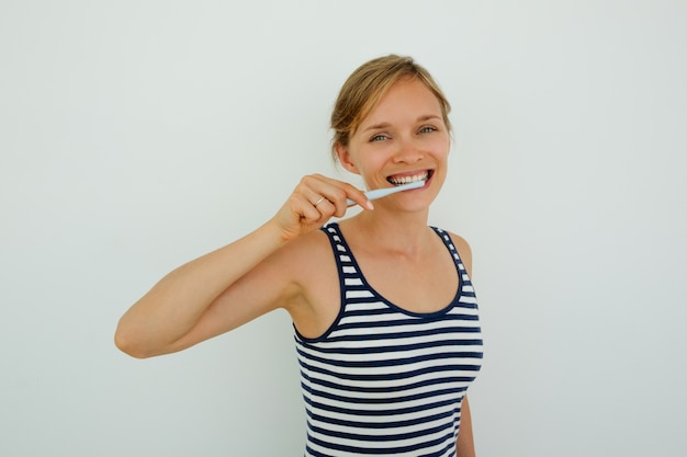 Cheerful young woman brushing teeth and looking at camera. Happy healthy girl scrubbing teeth with toothbrush equipped with toothpast. Oral hygiene routine concept