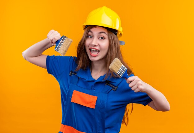 Cheerful young woman builder worker in construction uniform and safety helmet holding paint brushes smiling broadly 