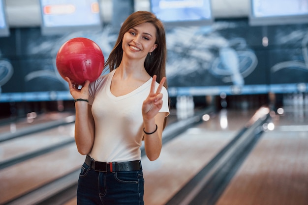 クラブで赤いボウリングのボールを保持しているカジュアルな服装で陽気な若い女性