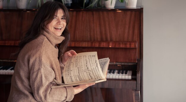 Free photo a cheerful young woman in a cozy sweater sits near the piano and looks at the notes.