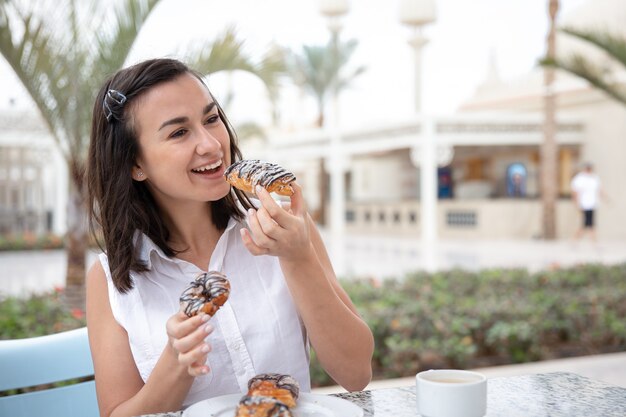 Cheerful young woman enjoying morning coffee with donuts on the outdoor terrace. Vacation and recreation concept.