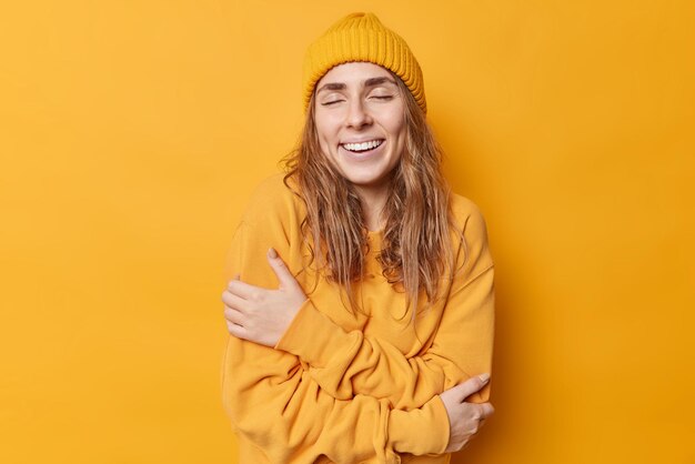 Cheerful young woman enjoys wearing comfortable jumper keeps eyes closed embraces own body expresses self love isolated over yellow background recalls something pleasant in mind Monochrome shot