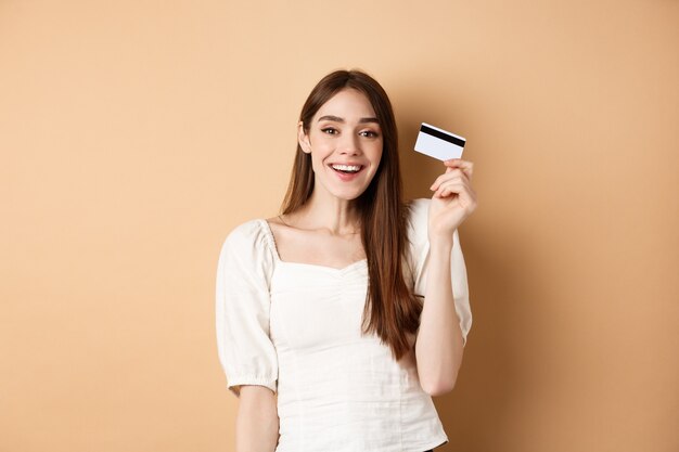 쾌활한 젊은 여성은 플라스틱 신용카드를 받고 베이지색 바탕에 만족스럽게 웃고 있습니다.