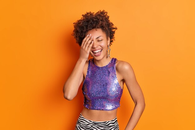 陽気な若い女性はアフロの髪の笑顔が広く顔に手を保ちます面白い話の上に楽しい笑いを感じますオレンジ色の壁に分離された紫色のトップスター型のイヤリングを着ています
