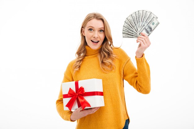 Cheerful young woman holding money and surprise gift box.