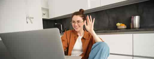 Бесплатное фото Веселая молодая женщина в очках говорит привет, махает рукой к ноутбуку, разговаривает с другом онлайн-видео