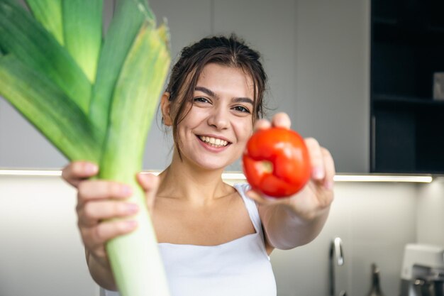 A cheerful young woman in the kitchen holds a leek and a tomato in her hands