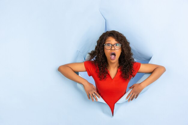 Cheerful young woman poses in torn blue paper hole background, emotional and expressive