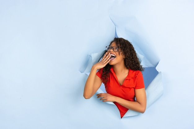 Cheerful young woman poses in torn blue paper hole background, emotional and expressive