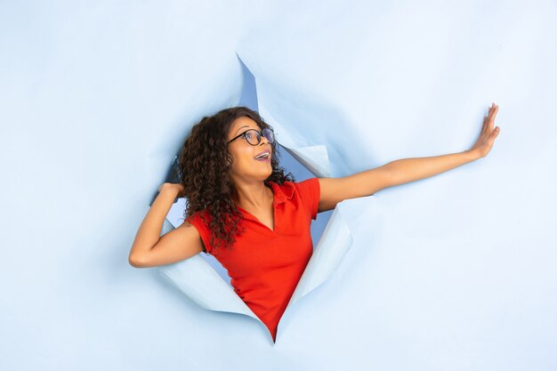 Cheerful young woman poses in torn blue paper hole background, emotional and expressive