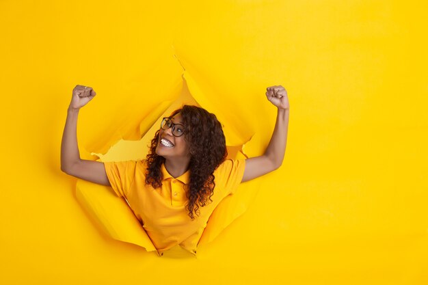 Cheerful young woman poses in torn yellow paper hole background, emotional and expressive