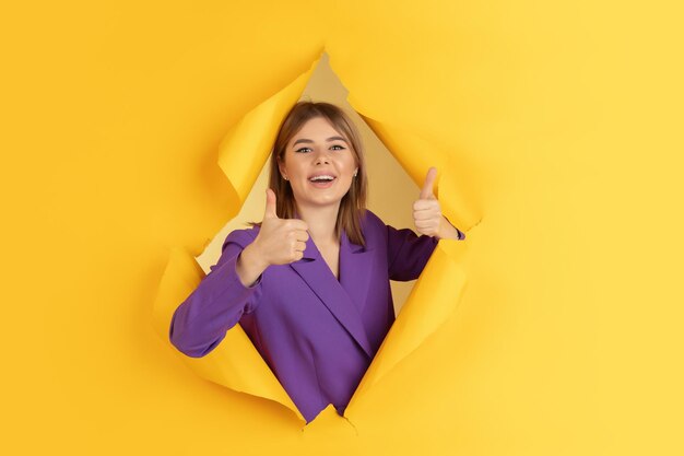 Cheerful young woman poses in torn yellow paper hole, emotional and expressive