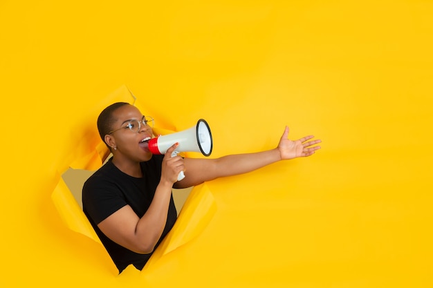 Free photo cheerful young woman poses in torn yellow paper hole wall emotional and expressive shouting and calling with speaker