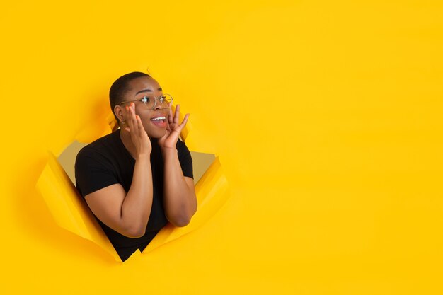 Cheerful young woman poses in torn yellow paper hole wall emotional and expressive shouting with speaker