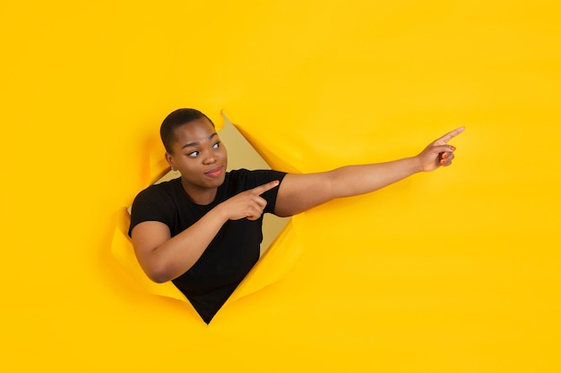 Free photo cheerful young woman poses in torn yellow paper hole wall emotional and expressive