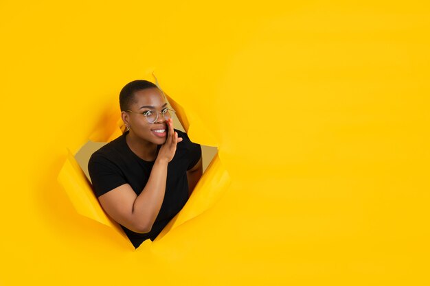 Cheerful young woman poses in torn yellow paper hole wall emotional and expressive