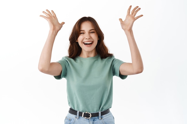 Cheerful young woman raising hands up, laughing and smiling carefree, winning and rejoicing, celebrating great news, standing over white wall