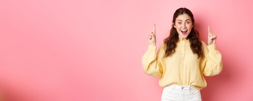 無料写真 広告を示す陽気な若い女性プロモーション テキストで指を上向きにし、ピンクの背景に対して興奮して立っている笑顔