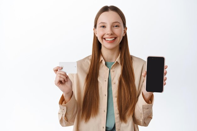 Cheerful young woman showing empty phone screen and credit card smiling pleased demonstrate mobile app for shopping standing over white background