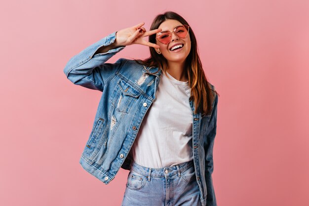 Cheerful young woman showing peace sign with smile. Studio shot of attractive caucasian lady wearing denim clothes.