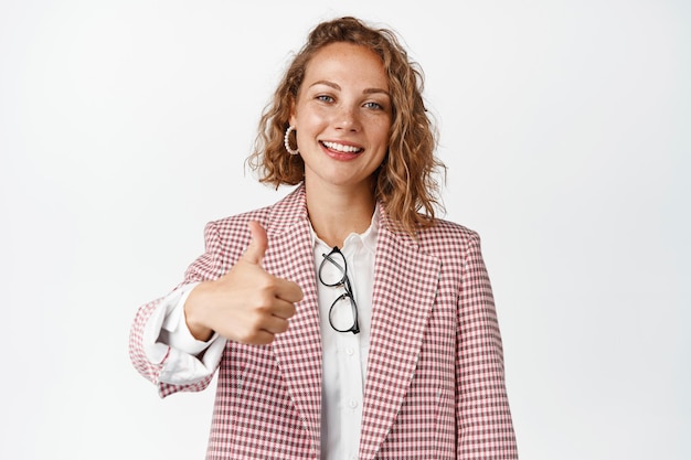 Cheerful young woman showing thumbs up, approve something good, smiling and nod, standing in suit against white background