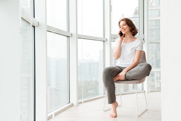 Cheerful young woman sitting near window talking by phone