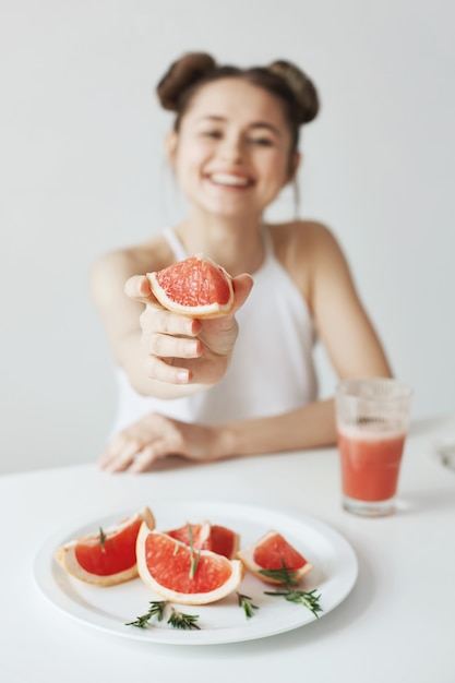 Cheerful young woman smiling sitting at table stretching piece of grapefruit over white wall. Healthy food concept.