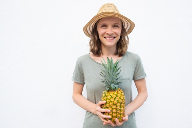 パイナップル全体を保持している麦わら帽子の陽気な若い女性