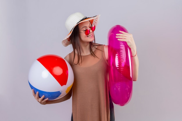Веселая молодая женщина в летней шляпе в красных солнцезащитных очках держит надувной мяч и улыбается, глядя на надувное кольцо стоя