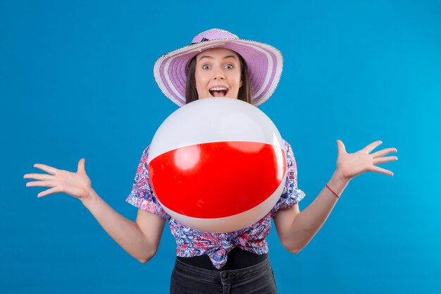 Cheerful young woman in summer hat wearing throwing inflatable ball smiling with happy face standing over blue space