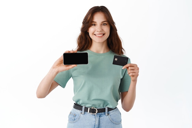 Tシャツを着た陽気な若い女性は、白い壁に立って、水平方向に空のスマートフォン画面とプラスチックのクレジットカードを示した