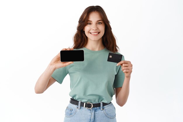 Веселая молодая женщина в футболке продемонстрировала пустой экран смартфона по горизонтали и пластиковую кредитную карту, стоя у белой стены