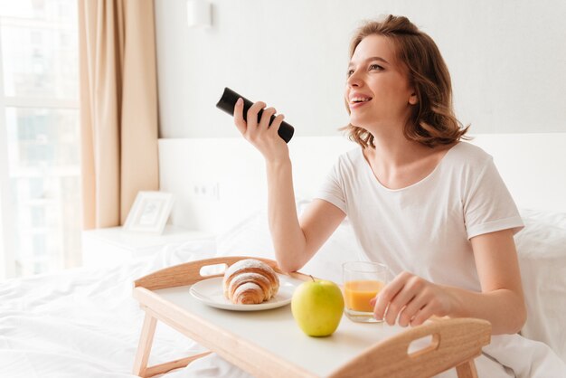 陽気な若い女性は、リモートコントロールを保持しているテレビを見ます。