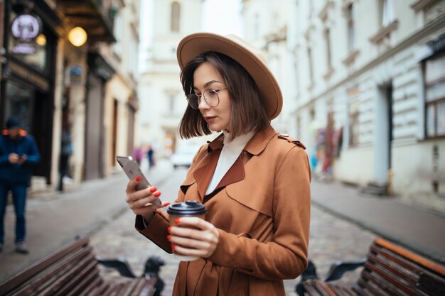 테이크 아웃 커피 컵을 들고 야외에서 걷는 코트를 입고 쾌활 한 젊은 여자