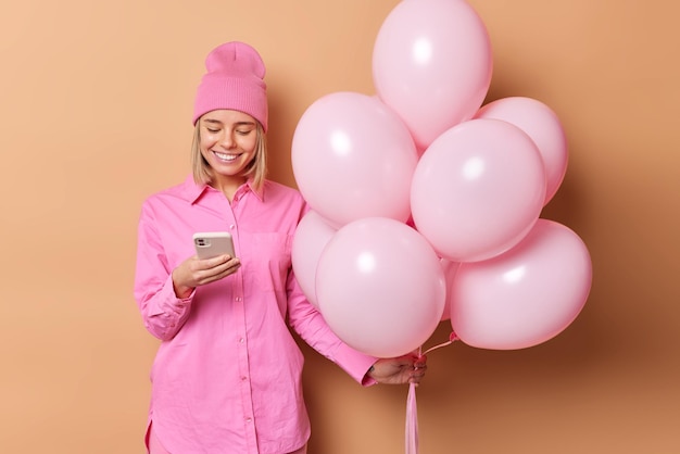 Cheerful young woman wears pink shirt and hat checks receieved message via smartphone holds bunch of inflated balloons celebrates special occasion isolated over brown background Holiday concept