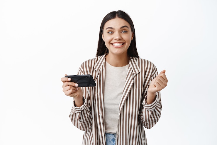 Free Photo cheerful young woman winning money on phone, achieve goal in smartphone app, holding mobile phone horizontally and say yes, white wall