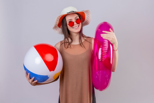 Веселая молодая женщина в шляпе в красных солнцезащитных очках держит надувной мяч и кольцо, улыбаясь, стоя на белом