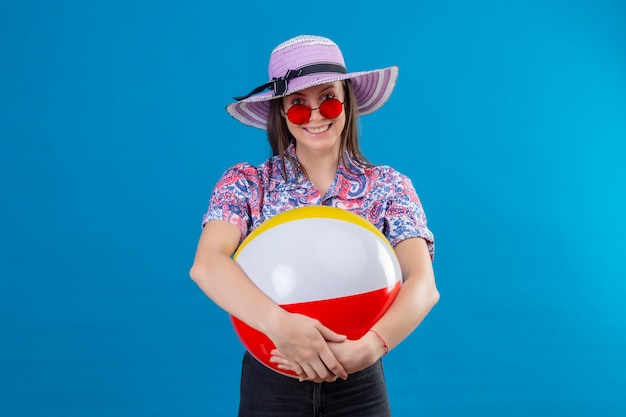 Cheerful young woman with hat wearing red sunglasses holding inflatable ball with smile on face standing on blue