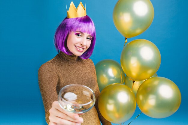 Giovane donna allegra con taglio di capelli viola che celebra la festa di capodanno con palloncini dorati e champagne. abito di lusso, corona in testa, compleanno, cocktail alcolico.